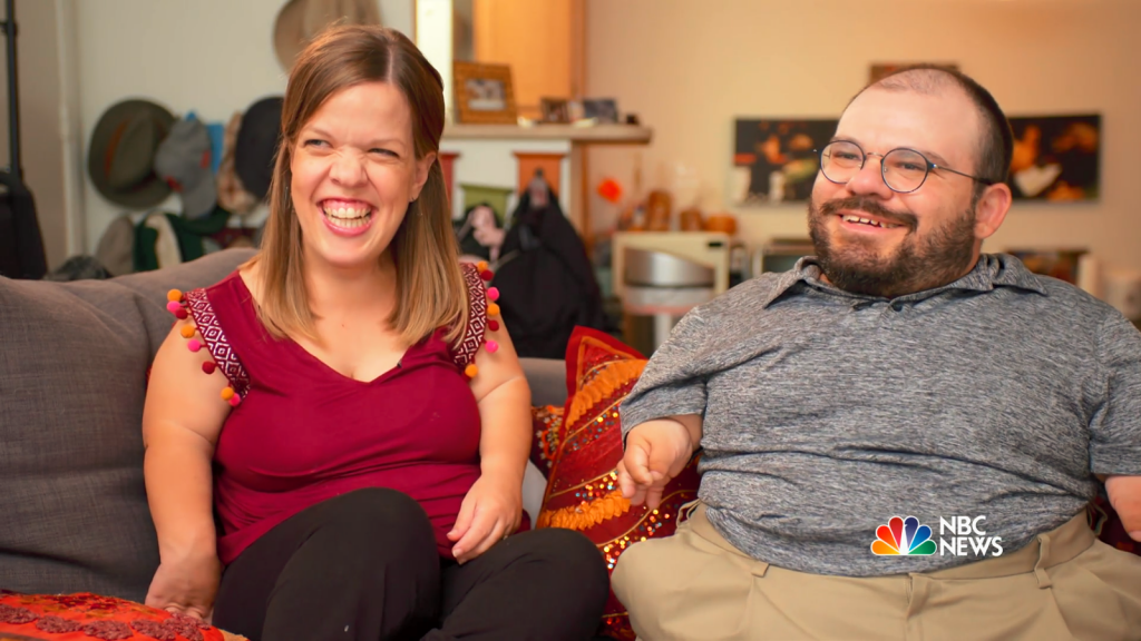 Leah Smith is smiling joyously, wearing a maroon top with orange and white accents, and black pants. Joe sits on the right, with glasses and a beard and small smile, wearing a grey polo shirt and khaki pants. Leah and Joe each have a different form of dwarfism.