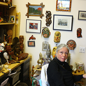 The wall behind her is covered with painted masks and framed pictures. Horizontal surfaces bear many statues made of wood and metal and ceramic. Dr. Silvers is looking attentively toward the photographer, her silver hair pulled back, wearing a black turtleneck with a necklace.
