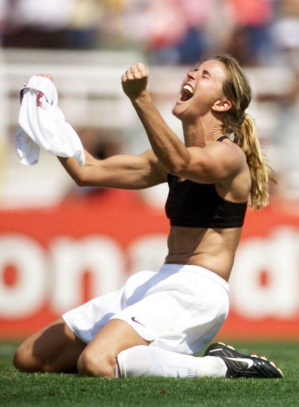 Brandi Chastain, skin pale, kneels on the ground, shirt in her hand, sports bra and belly exposed to view, arms up-raised with every muscle tensed, head back and shouting 