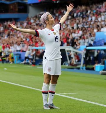 Megan Rapinoe, a muscular but lean white athlete with bleached blonde hair died lavender, stands with her feet planted solidly and her arms outstretched, smiling, after scoring a game-changing goal against a difficult opponent..