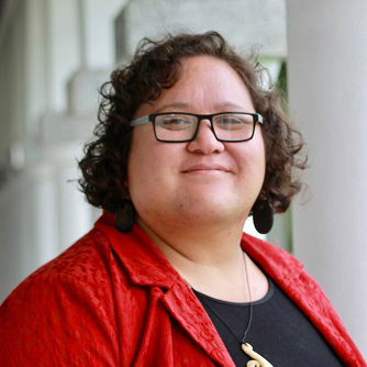 A sturdy woman with jaw-length curly brown hair and rectangular black glasses smilles slightly toward the camera.  She is wearing a bright red blazer and black shirt. Her necklace appears to be traditional Māori carved bone art.