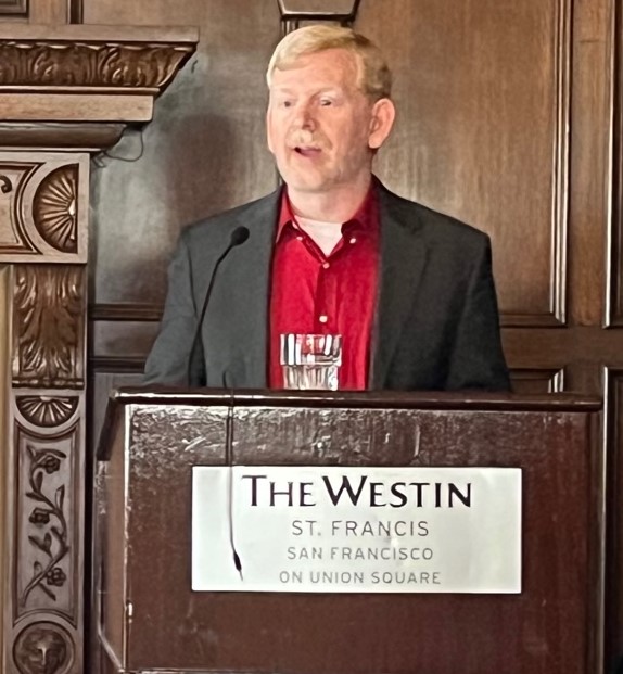 Adam Cureton, who has short neatly trimmed blonde hair and is wearing a bright red shirt with a grey suit jacket, stands at the podium speaking