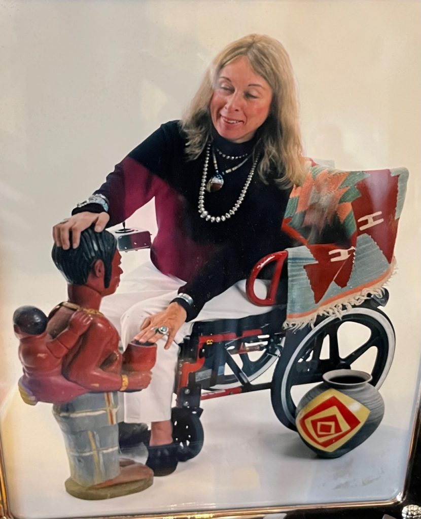 Anita Silvers is depicted. She is sitting in  her powerchair, which has snazzy black and red detailing. She has pale skin, long blonde hair that is greying, is wearing white slacks and a black turtleneck, and wearing large beaded silver jewelry including necklaces and a turqoise ring. She has a blanket across the back of her chair colorfully decorated in geometric patterns common in North American indigenous peoples' blankets. She is gazing at and touching a ceramic sculpture of a mesoamerican person wearing an infant on their back. A ceramic pot rests on the floor next to the left wheel of her chair.