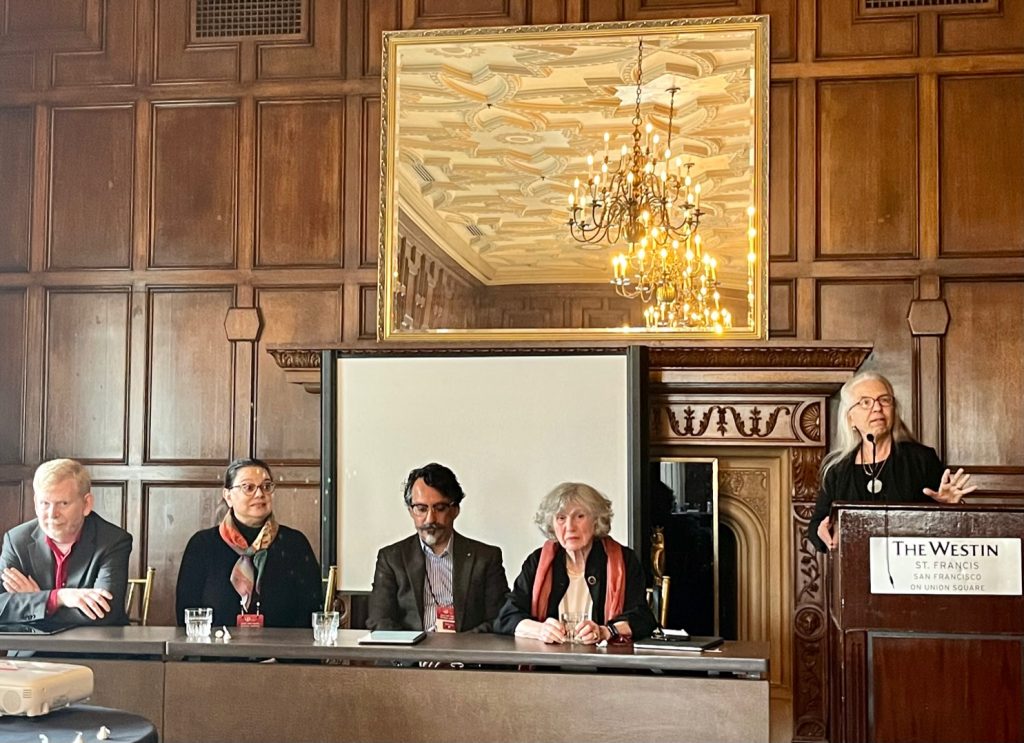 The full panel sits at a long table in the front of a conference room. There are two light-skinned women and two light-skinned men at the table, all paying attention to a speaker who is standing at a podium next to them gesturing animatedly with her hand. Behind them is an ornate wood-paneled wall and carved fireplace with mantle, as well as a mirror reflecting the warm yellow light of chandeliers.