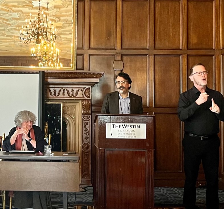 Dr. Dominic McIver Lopes stands at the podium speaking. He has light skin, very dark wavy ear-length hair and mustache, and a small, neatly trimmed grey goatee. To his right is an ASL interpreter. Sitting at the panel table to his left is Eva Kittay, listening attentively.