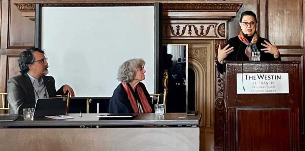 Teresa Blankemeyer-Burke stands at the podium speaking animatedly with both her voice and her hands. She has long dark hair pulled back into a ponytail, a black turtleneck shirt, and a colorful scarf around her neck. To her left, turned to face her attentively, are Dr. Dominic McIvers Lopes and Dr. Eva Kittay. Behind her is the same wood-paneled background and carved fireplace mantle as in previous photos of the conference session.